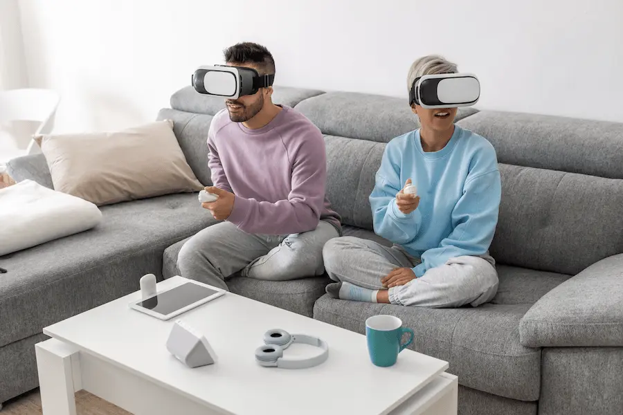 Couple enjoying gaming with AR gadgets, wearing virtual reality headsets while sitting on a sofa in their living room.