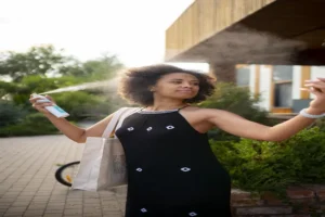 Heat Protectant Spray A woman in a black dress is spraying mist from a bottle on her face while standing outdoors. She carries a canvas tote bag on her shoulder and appears relaxed and refreshed, using what could be the Best-Heat-Protectant-Spray to stay cool and protect her skin or hair from heat