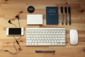 An gadget Apple setup with an iPhone, wireless keyboard, mouse, notebook, and accessories including Canon lens, external hard drive, and pens, perfect for a tech enthusiast's workspace.