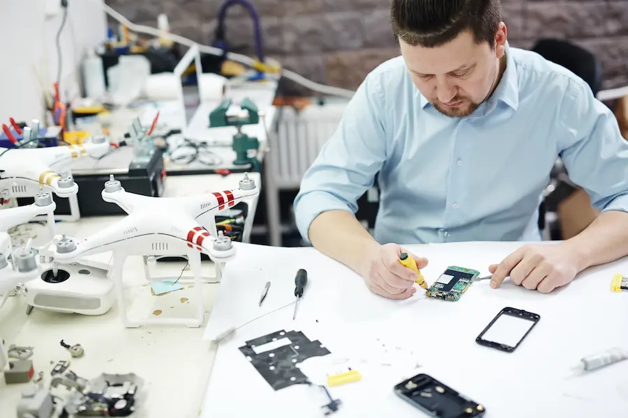 Technician performing gadget repair on smartphone motherboard at a tech workbench with drones and tools