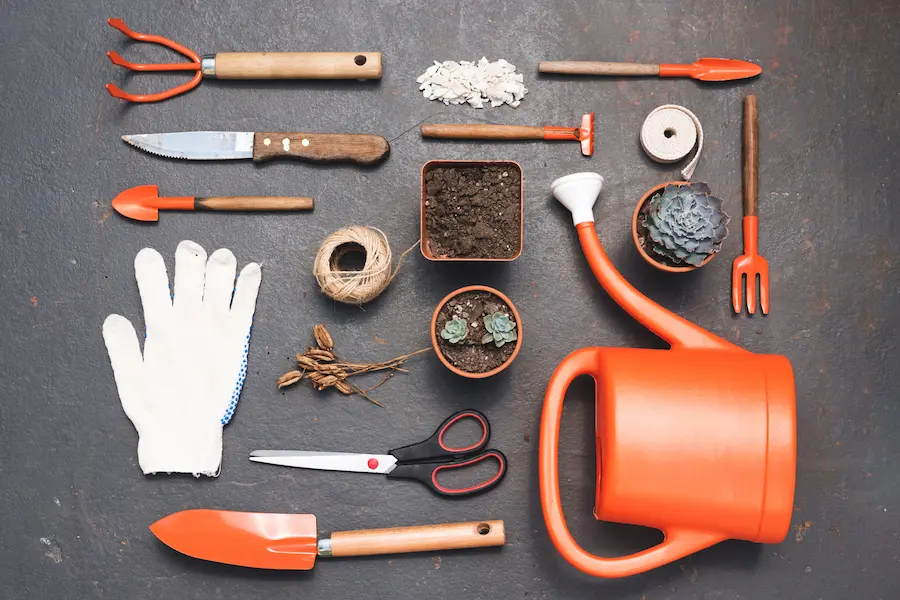 A collection of essential garden gadgets, including a watering can, gloves, trowels, scissors, and small pots, arranged neatly on a dark surface.